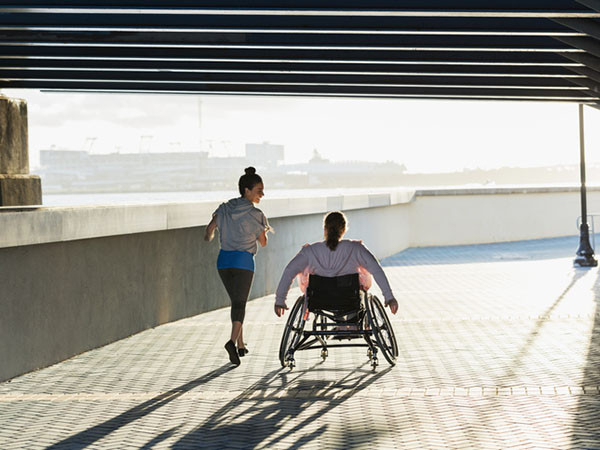 view from behind of two women exercising along a city waterfront, passing under a bridge, woman on the left is jogging while woman on the right is using a wheelchair